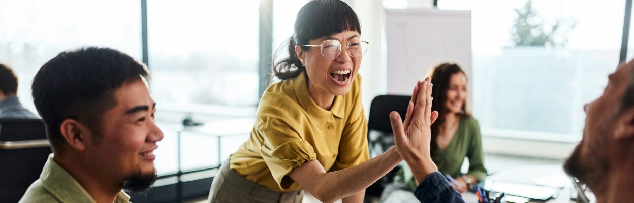 A group of four colleagues cheering and smiling in an office. Two of the colleagues are giving each other a high five.